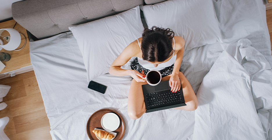 Brunette lady in summer pajamas enjoying coffee and working on a laptop whilst sitting in a bed with light grey bedding and grey headboard 